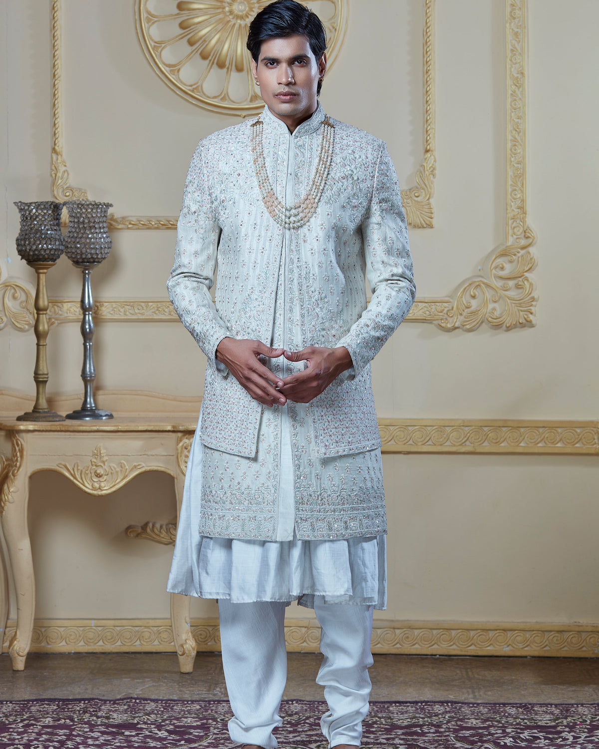 A man modeling a traditional white Indian kurta set with intricate silver embroidery, paired with white trousers and brown shoes, standing in an ornate cream-colored room with gold wall details | Aditya Bhardwaj