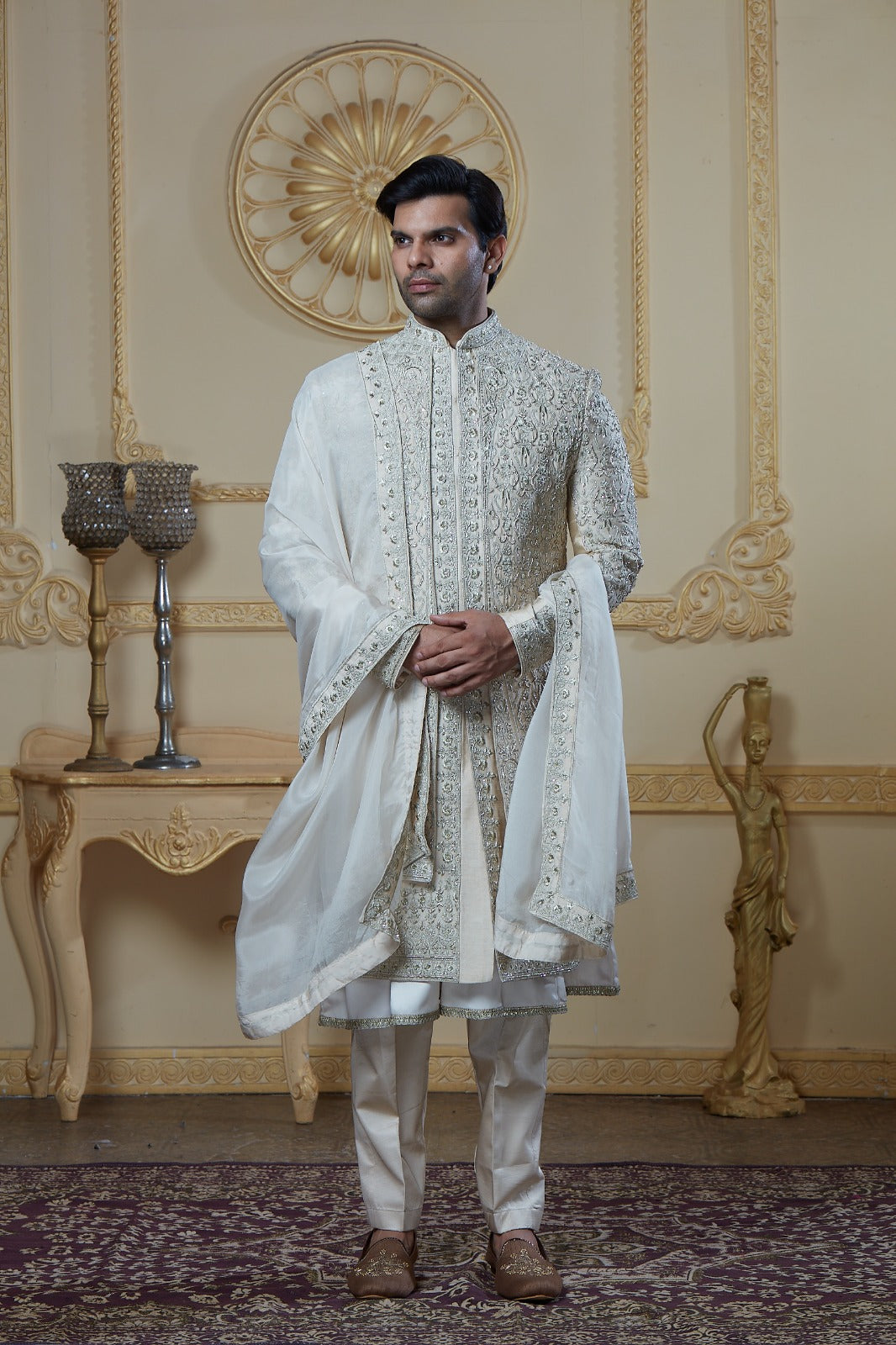 A man modeling a traditional white Indian kurta set with intricate silver embroidery, paired with white trousers and brown shoes, standing in an ornate cream-colored room with gold wall details | Aditya Bhardwaj