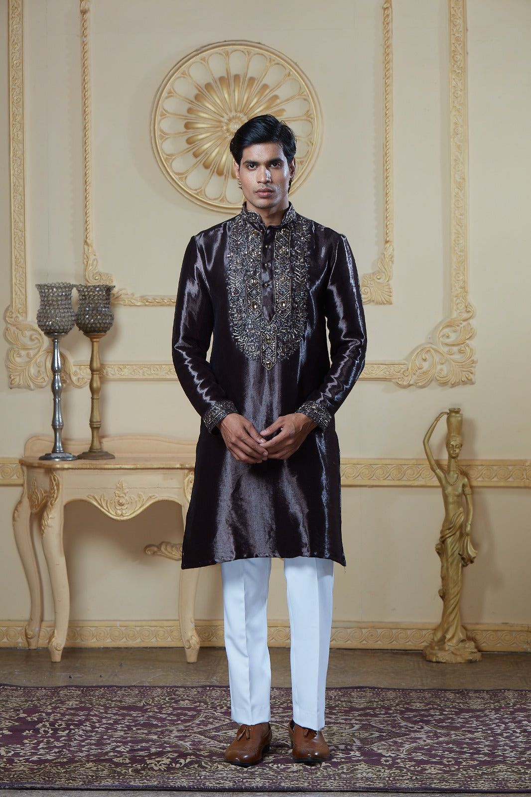 A man modeling a dark brown metallic kurta with intricate silver embroidery on the chest and collar, paired with white trousers and brown shoes, standing in an ornate cream-colored room | Aditya Bhardwaj