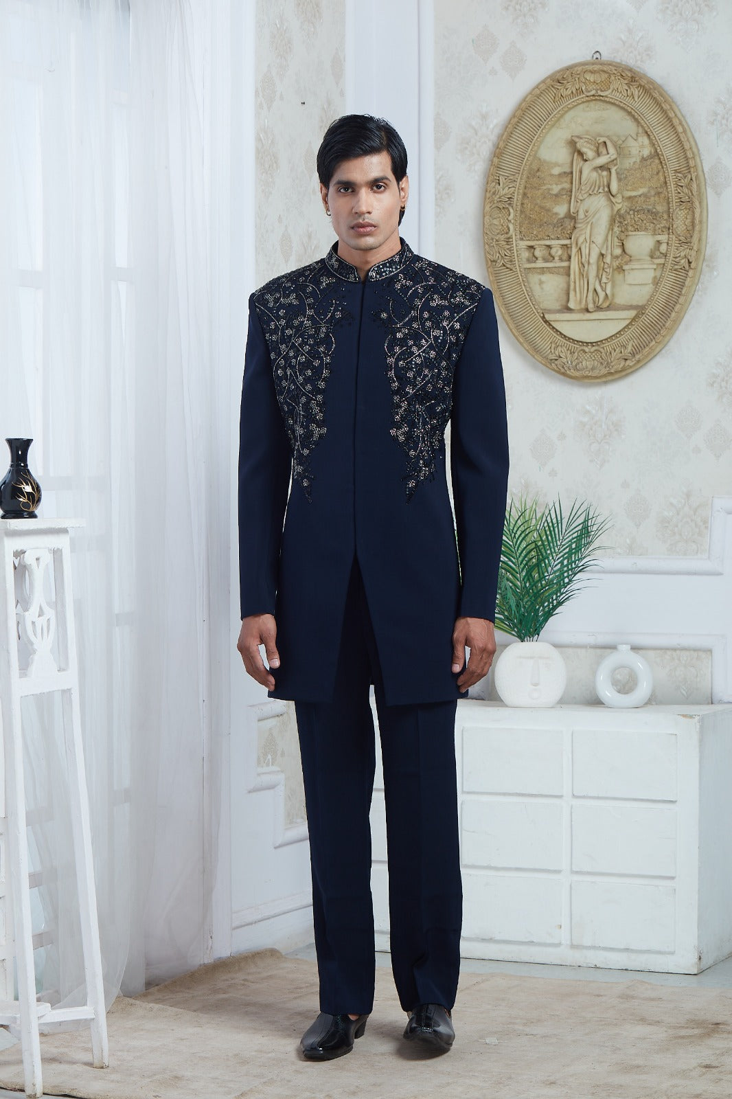 A man wearing a dark navy blue traditional Indian suit with intricate silver embroidery on the chest, standing in a decorated room with white walls and furniture | Aditya Bhardwaj