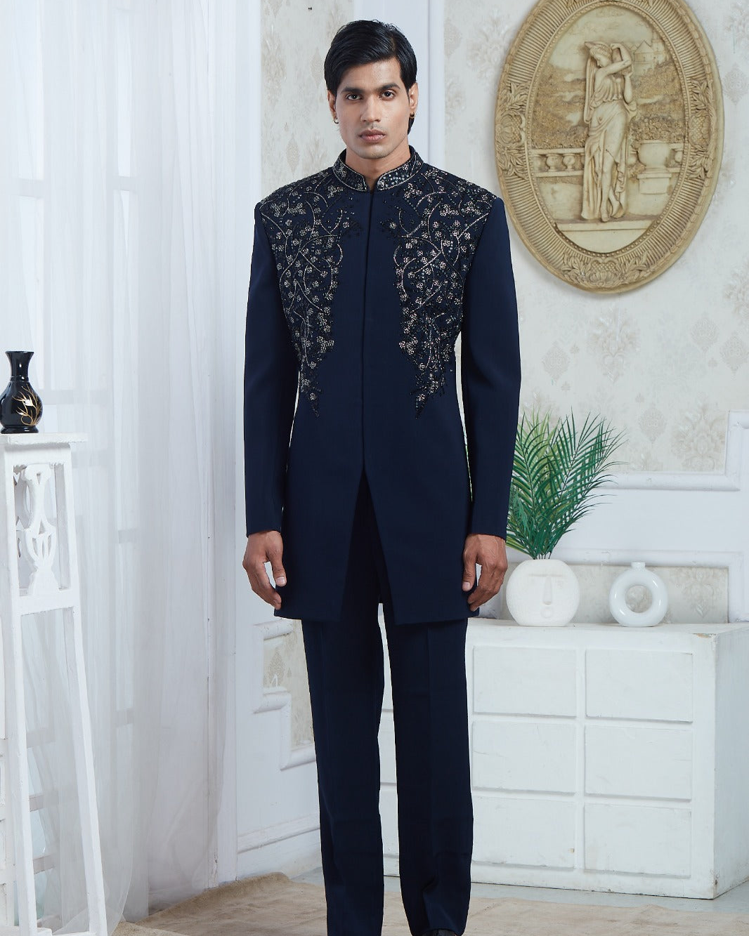 A man wearing a dark navy blue traditional Indian suit with intricate silver embroidery on the chest, standing in a decorated room with white walls and furniture | Aditya Bhardwaj