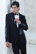 A man wearing a black tuxedo jacket with silver sequin embellishments on the lapels and sleeves, paired with a white dress shirt and black bow tie, standing in an elegant room with white walls and patterned wallpaper | Aditya Bhardwaj