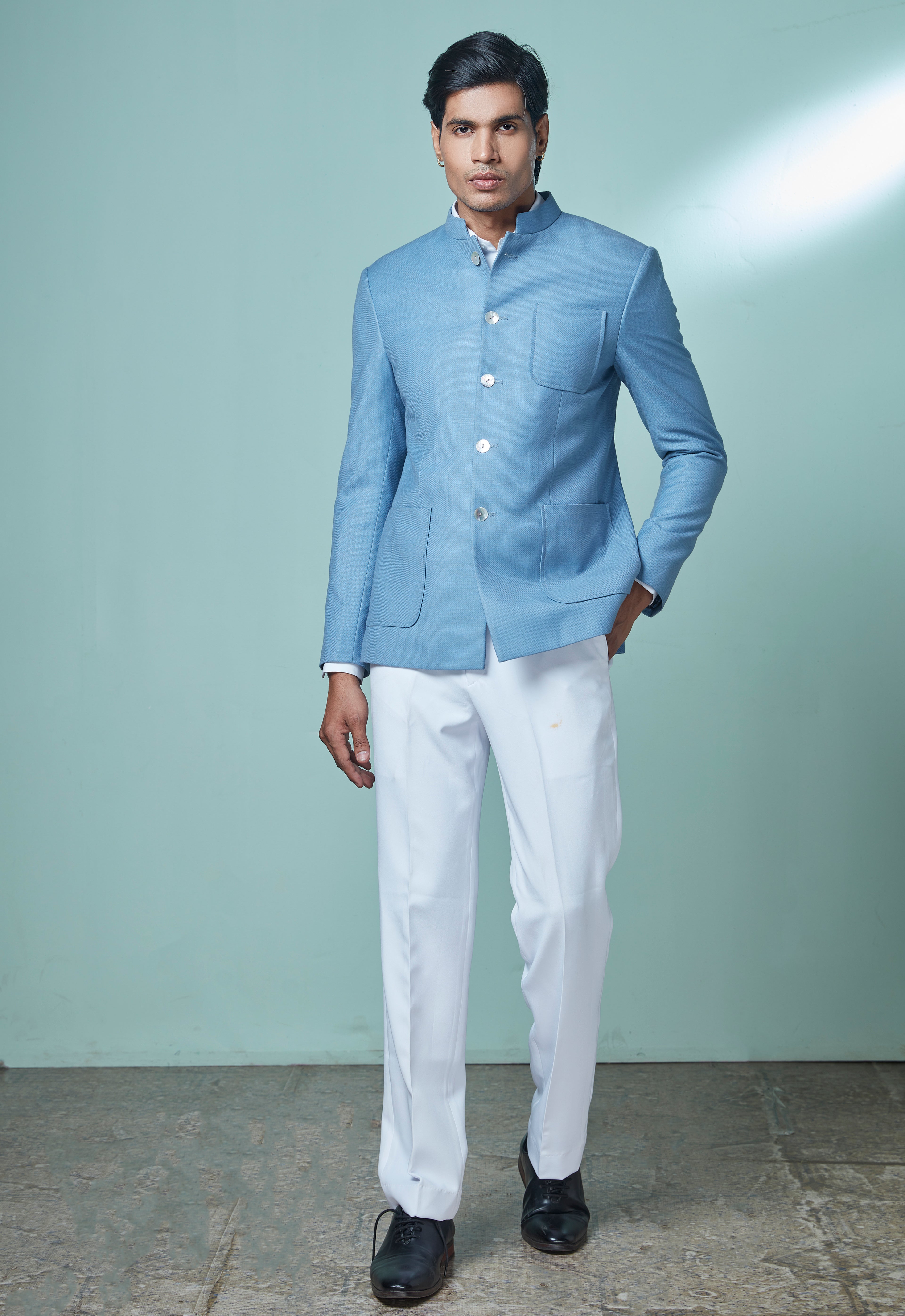 A man modeling a light blue Indian ethnic jacket (kurta) paired with white trousers and black shoes, standing against a light green studio backdrop.