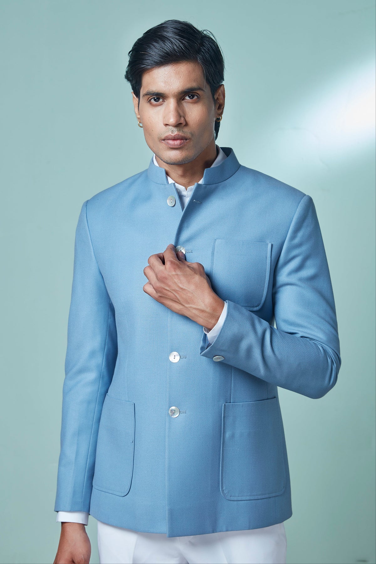 A man wearing a light blue Indian mandarin collar blazer (kurta) with white trousers, standing against a solid light green studio background.